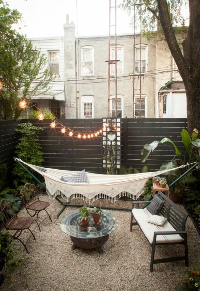 como decorar una terraza pequeña, terraza con hamaca blanca, mesa redonda de vidrio, suelo de gravilla, bombillas colgantes