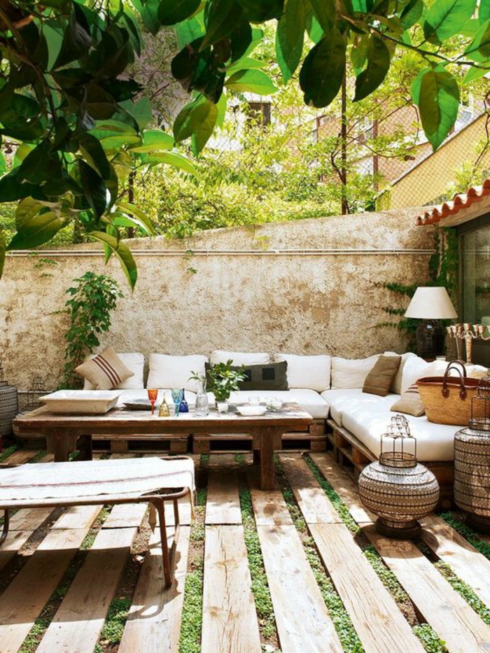 decoracion jardin, terraza con suelo de madera y césped, sofá con cojines blancos, mesa con vasos y lámpara