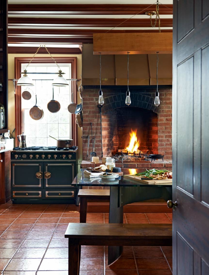 cocinas rusticas, cocina con chimenea de ladrillo encendido, sartnes colgantes, mesa y suelo de baldosas