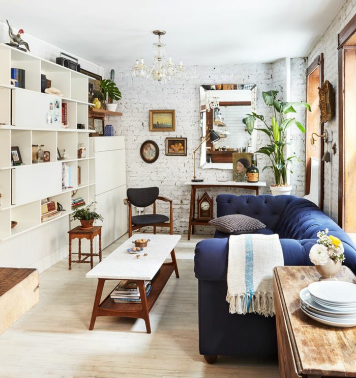 decoracion salones pequeños, salón con estantería blanca y sofá azul, pared de ladrillo y lámpara de araña, mucha luz natural, plantas verdes