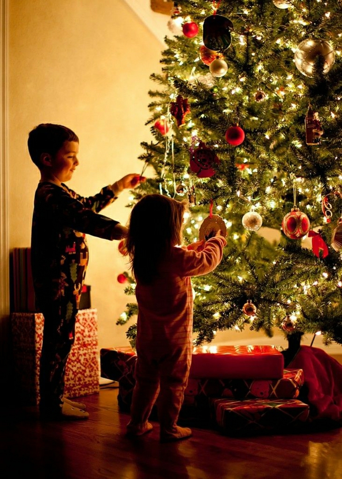 arbol de navidad decorado, pino grande con decoración en los colores clásicos para navidad, niños colocando los adornos