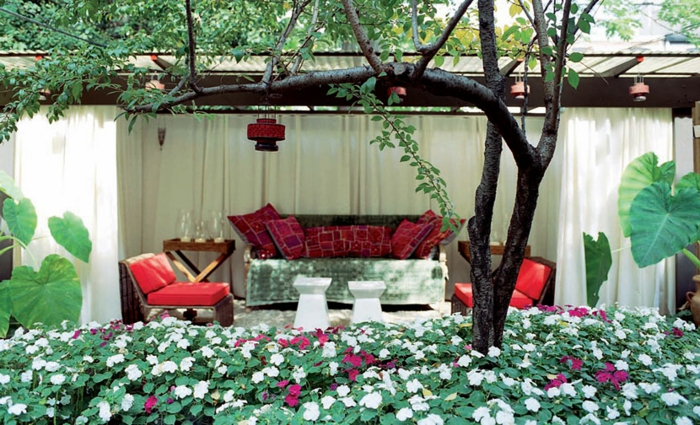 jardines con piedras, veranda original con muebles sacados fuera, colchonetas y cojines en rojo, muchas plantas con flores blancos