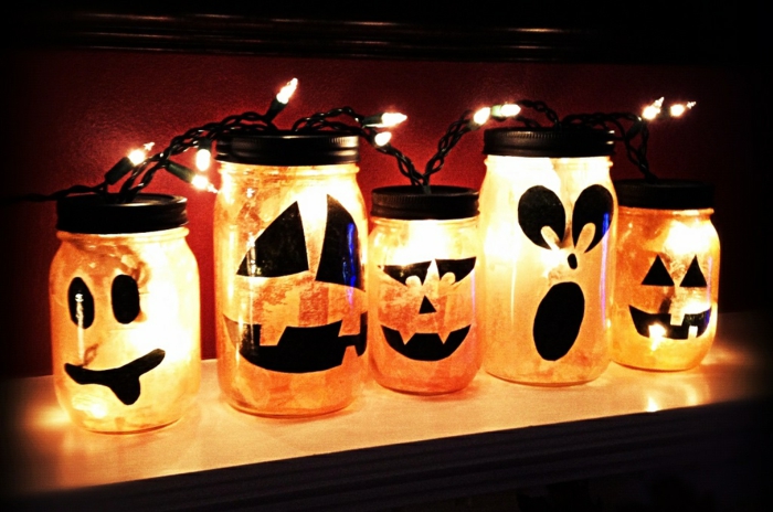 manualidades caseros, frascos con caras pintadas en negro, bombillas y velas,puestos en un estante de madera