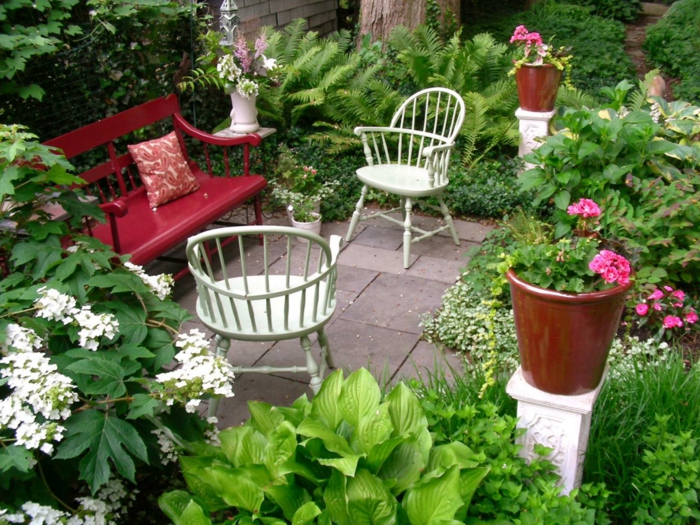como hacer un jardín en un terreno pequeño, banco rojo de madera con sillas blancas vintage, macetas con geranios, muchos arbustos de diferente tipo