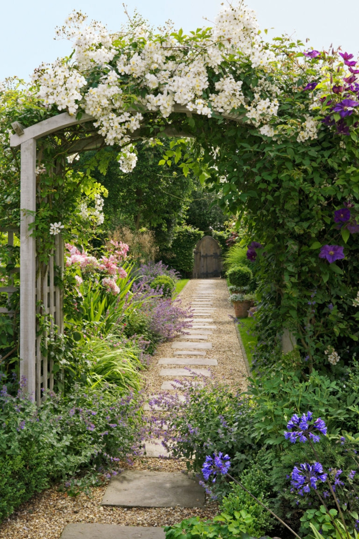 diseño de jardines, bonita bóveda con plantas enredaderas en blanco y lila, sendero con pavimento de piedras, seto vivo alto, jardín con toque salvaje
