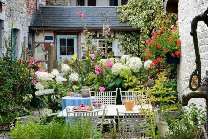 jardines con piedras, patio estilo provenzal colorido, con grandes flores, sillas y mesa de madera blancos, mucha decoración