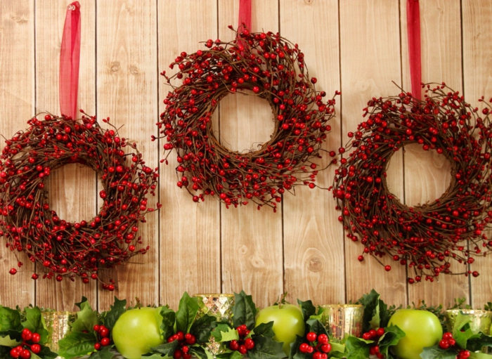 adornos de navidad caseros, tres coronas navideñas de muerdago rojo con cintas rojas, guirnalda con hojas verdes y manzanas