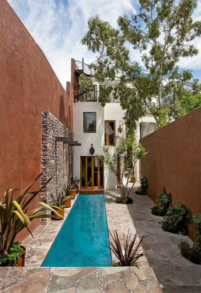 piscina desmontable, casa moderna con patio, suelo de piedras, piscina azul estrecha y larga, muros altos y árbol