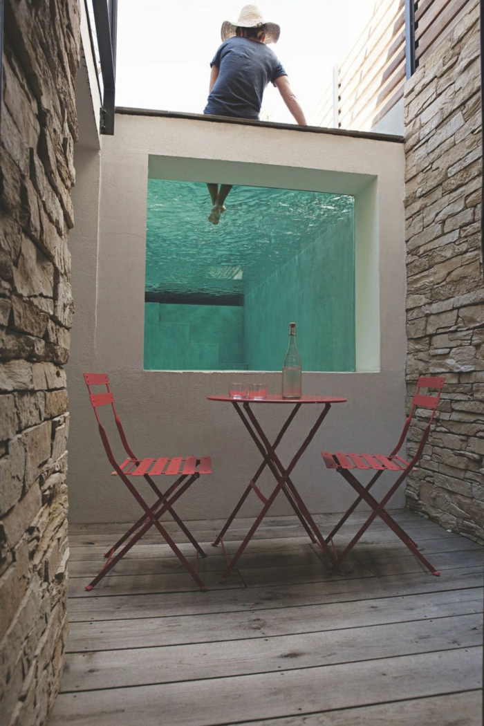 piscinas desmontables, piscina alta entre muros de piedra, hombre con sombrero sentado, mesa y sillas rojas, botella de vidrio