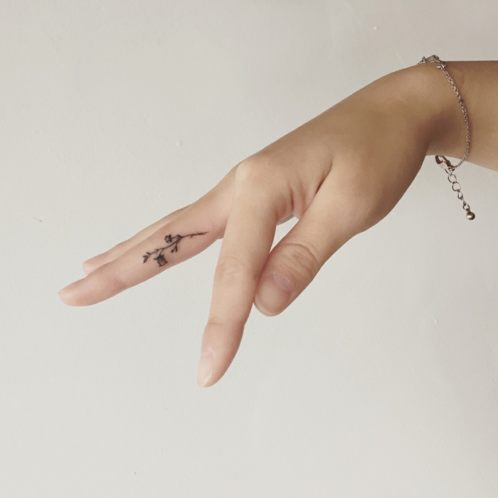 tatuajes mujer, mano delgada de mujer con pulsera de plata, tatuaje minimalista con flor en el dedo corazón