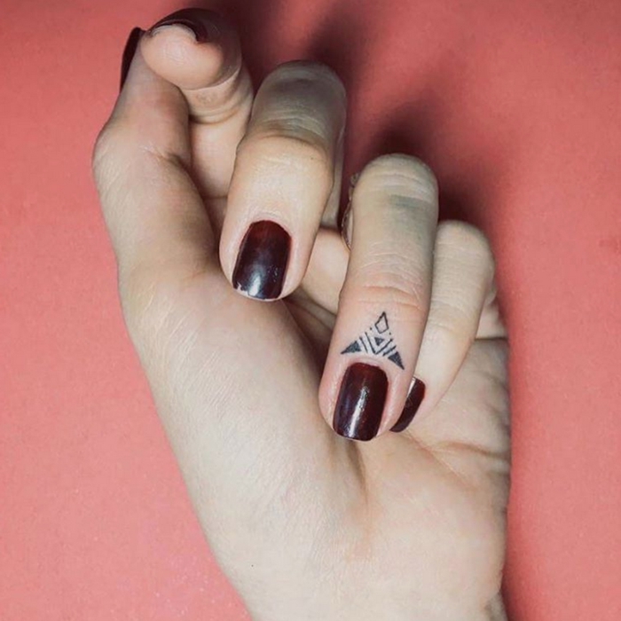tatuajes en los dedos, mano de mujer con pequeño tatuaje en forma de triángulo en el anular, uñas con esmalte carmín