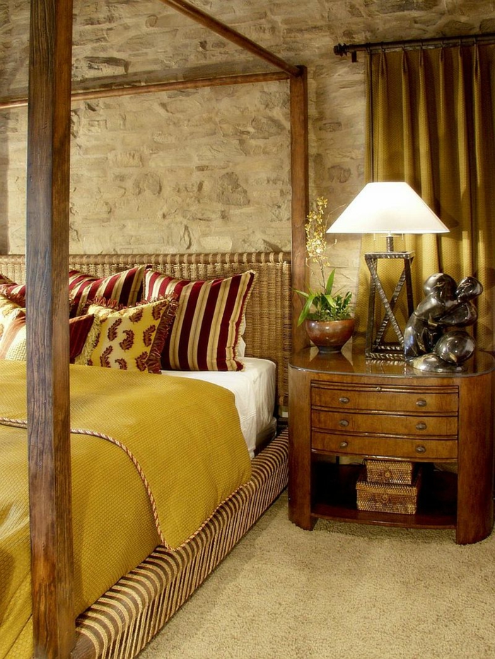 pared ladrillo, dormitorio en rojo y dorado, cama doble con pilares, mesita de noche de madera con estatua, pared de piedra rústica