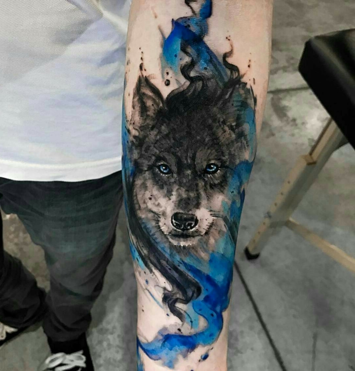 tatuajes de lobos, hombre con tatuaje en el antebrazo, tatuaje en azul y negro estilo acuarela, cabeza de lobo con ojos azules