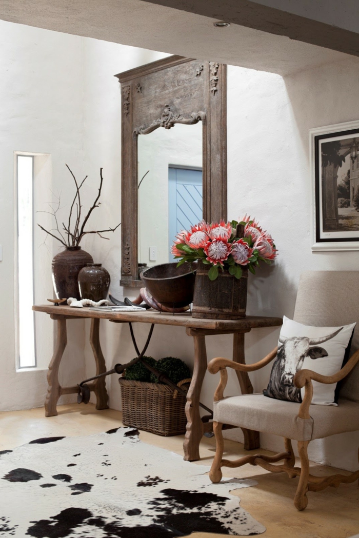 recibidores baratos, idea de decoración de recibidor en estilo rústico, alfombra piel de vaca, sillón con cojín, espejo grande antiguo, mesa auxiliar de madera con flores