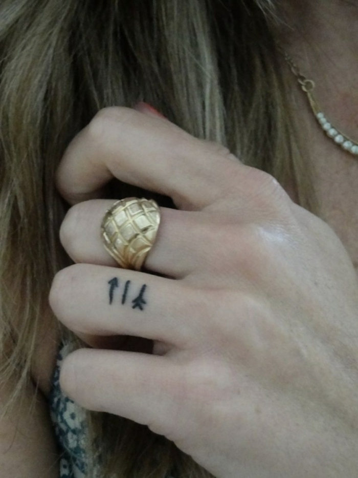 tatuaje corazon, mujer con pelo largo, tatuaje minimalista con flechas negras en el dedo anular, anillo de oro