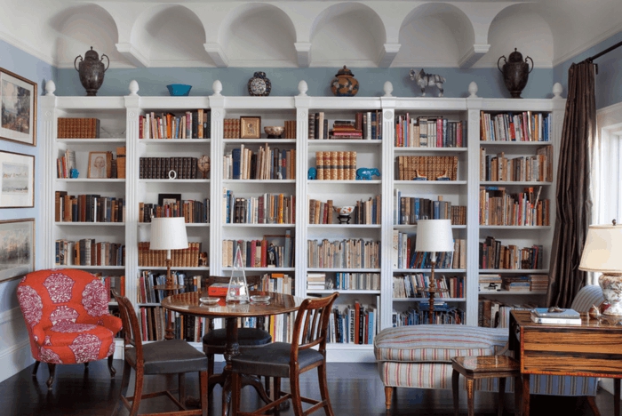 librería clásica de madera blanca, baldas de madera, salón con sillón rojo y mesa redonda, paredes en blanco y azul celeste, decoración con jarrones de barro