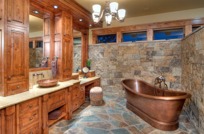 azulejos para baños que imitan piedra, baño en estilo rústico con bañera exenta vintage de cobre y muchos armarios de madera, candelabro vintage