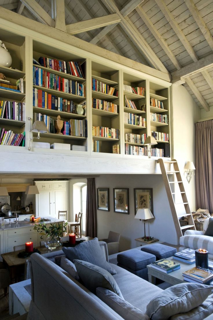 librerias, cocina abierta al salón, balcón interior con biblioteca, escalera de mano, techo triangular con vigas de madera, sofá azul