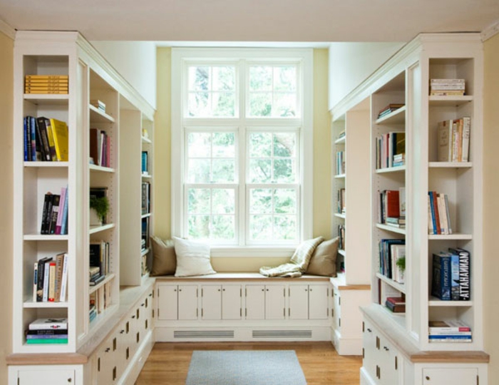 librerias, rincón de lectura cerca de la ventana, armario banco con cojines, estanterías de madera blanca, suelo con parquet y tapete azul