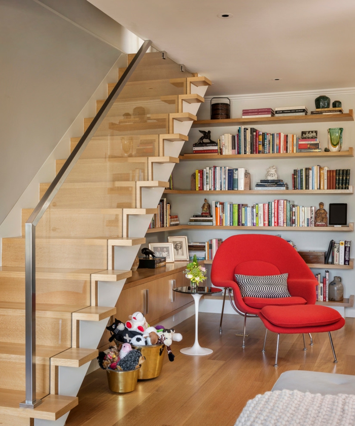 rincón de lectura bjo una escalera con barandilla de vidrio, estantes de madera con libros y decoraciones, sillón rojo con cojin an blanco y negro, librerías