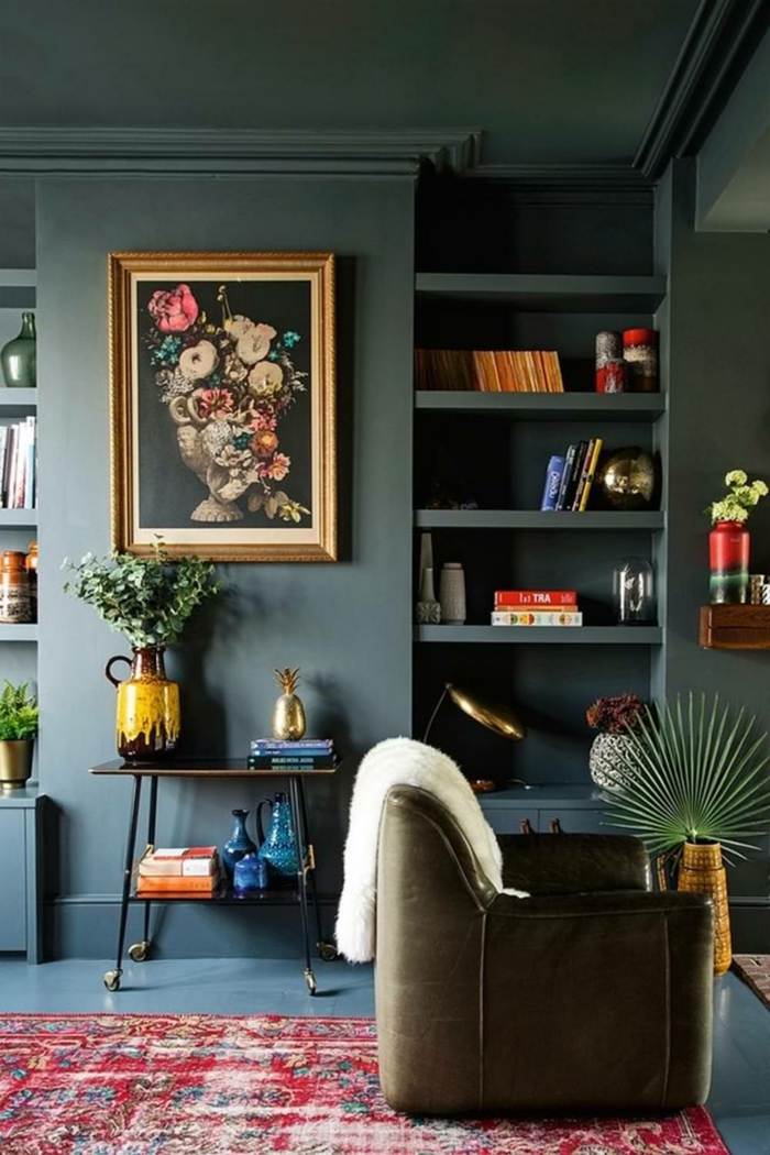 decoración en colores oscuros, colores para salones, salon pequeño, paredes azul oscuro grisáceo, cuadro con flores, sillon de pile, alfombra roja, estantería con libros