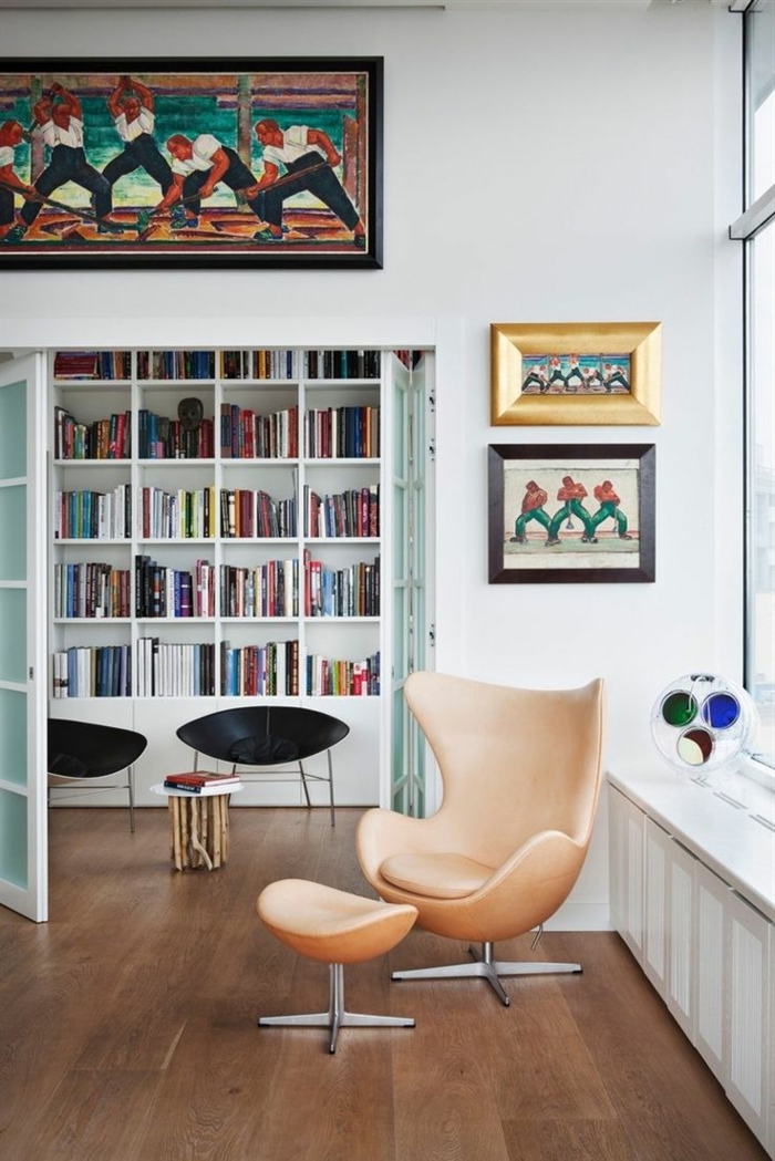 librerias, idea de ecoración con librería grande de madera blanca, puertas con vidrio, sillón moderno tapizado con taburete, cuadros con obreros