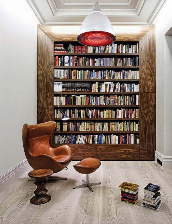 librerias, rincón de lectura con sillón tapizado de piel y taburete, librería empotrada con marco grande de madera, suelo con tarima, lámpara colgante roja de metal y vidrio