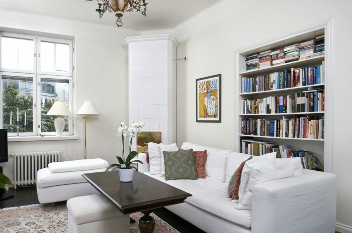 salón decorado en blanco, librería empotrada, mesa de madera con orquidea blanca, suelo laminado con alfombra, ventana pequeña, lámpara de araña, librerías