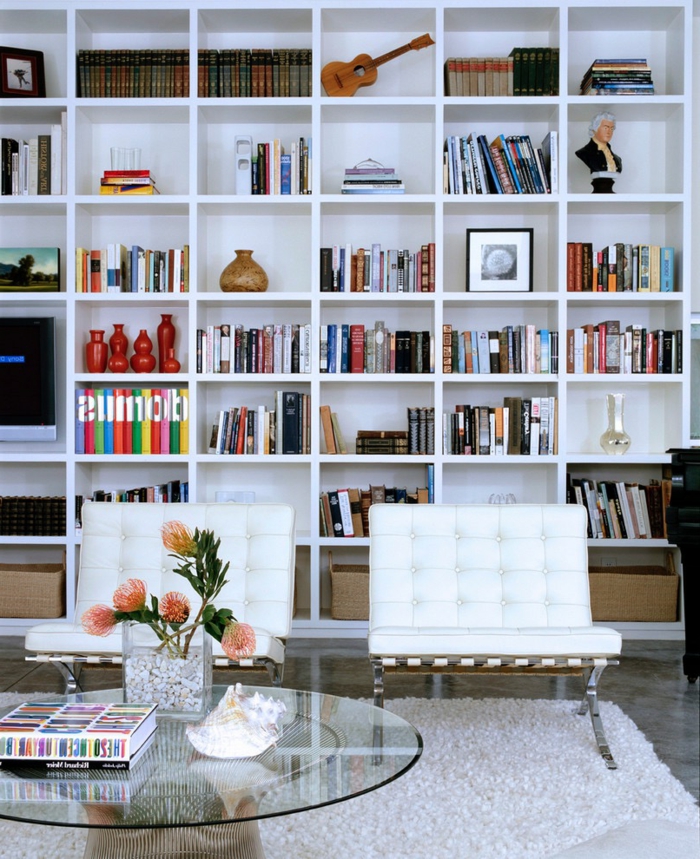 salón con librería de madera blanca, sillones tapizados en capitoné, mesa redonda de vidrio con flores, librerias, decoraciones con jarrones, estatuillas