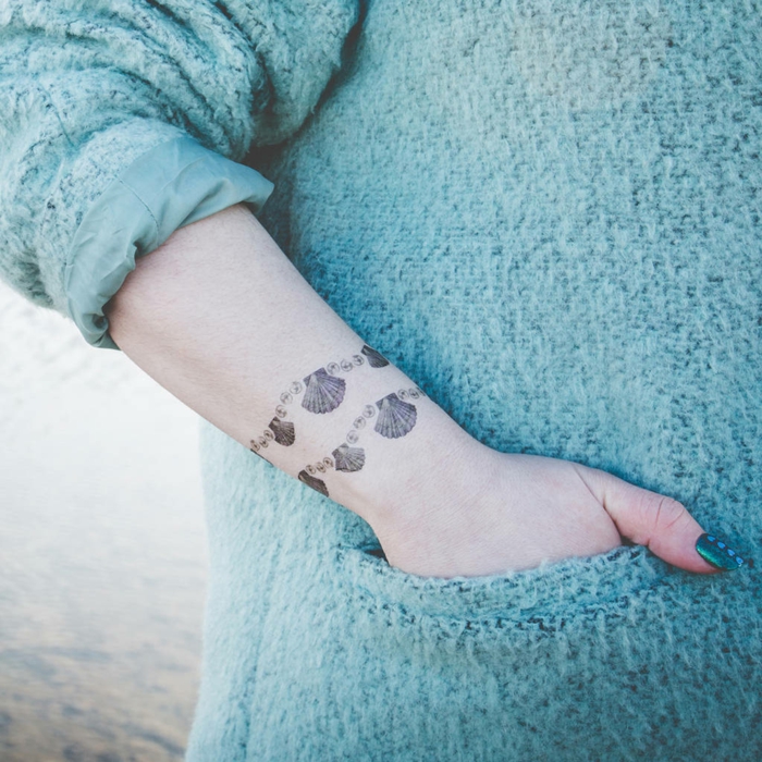 mujer con jersey aguamarina, tatuaje brazalete en la muñeca con cocnchas de mar en dos, uñas pintadas de verde metálico, tatuajes originales