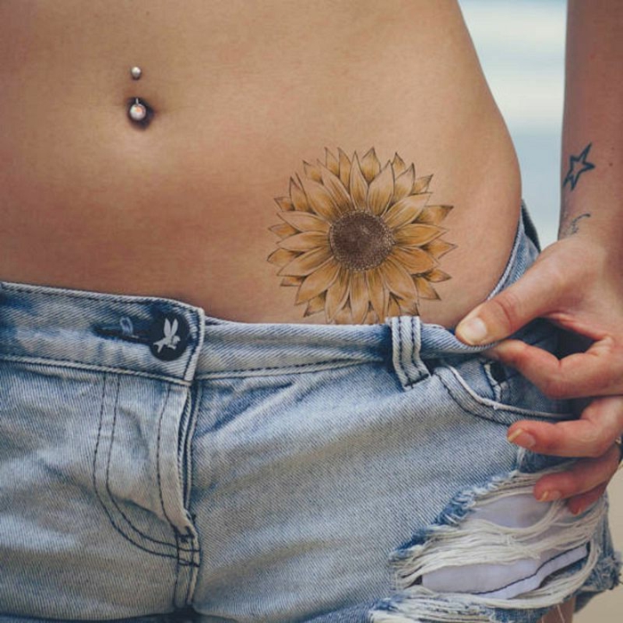 tattoos pequeños, mujer con piercing en el ombligo, tatuaje de girasol de color en el pelvis, jeans cortos