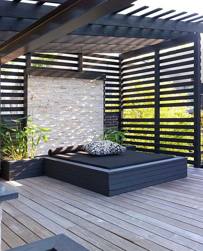 diseño sencillo y moderno de una terraza con grande cama en negro, suelo de madera, pérgola moderna pintada en negro 