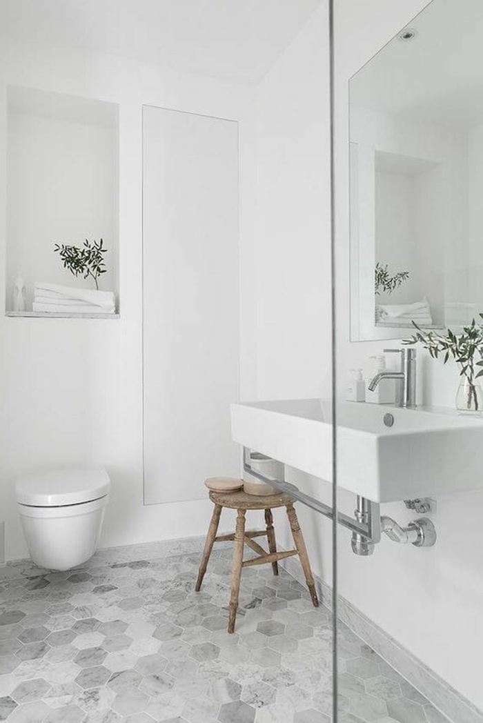 decoracion en blanco y gris, suelo con baldosas hexagonales, espejo grande, taburete de madera rustico, baños pequeños con ducha, ramas con hojas verdes
