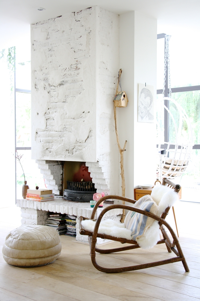 decoracion estilo nordico, salón con luz natural, chimenea de obra con ladrillo visto, pintura blanca, sillón de bambú con cojín, puf de cuero, casa para pájaros