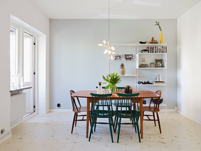 muebles estilo nordico, comedor con pocos muebles, mesa de madera, sillas en marrón y verde, estantería blanca alta de metal, lámpara colgante, suelo con tarima, ventanas sin cortinas