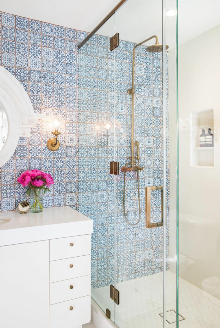 baño en estilo ecléctico, pared con azulejos en azul, ducha con puerta de vidrio y asa dorada, espejo redondo, mueble lavabo grande con flores ciclamen