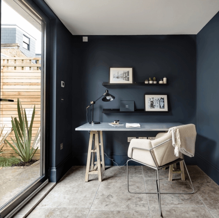 decoracion nordica salon, despacho pequeño con ventanal y terraza, paredes en azul oscuro, mesa con piernas de madera y encimera de plástico, silla tapizada con cuero, cuadros enmarcados