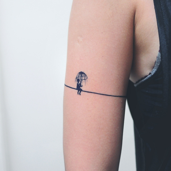 tatuajes pequeños, brazo mujer delgada, tatuaje tipo brazalete con niña bajo la lluvia con paraguas, color azul oscuro