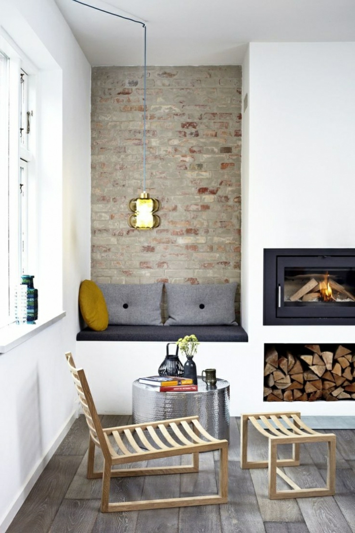 habitación pequeña, chimenea encendida, muebles estilo nordico, nicho almacenamiento de leña, pared de ladrillo visto, rincón de lectura, sillón con taburete de madera, mesa redonda baja