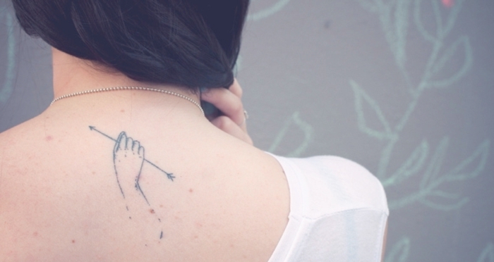 tatuaje en la espalda, mujer con blusa blanca y lunares, tatuajes chicos, mano colgando un flecho, pelo largo a un lado