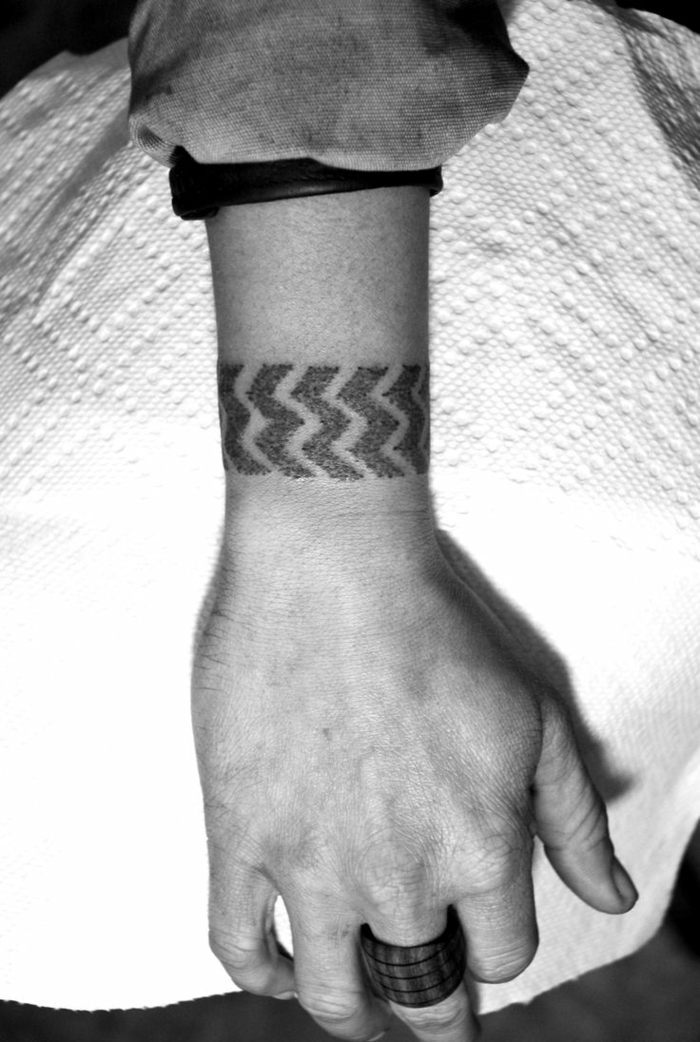 diseño de tatuaje brazalete masculino con líneas negras en la muñeca, foto en blanco y negro, mano con anillo grande, tattoos pequeños