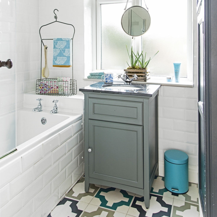 ducha con bañera, ventana mate, espejo colgante redondo pequeño, suelos con baldosas geométricas, armario bajo de madera pintada gris, cubo de basura azul