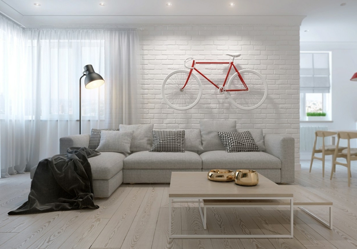 estilo nórdico, sala de estar, ladrillo visto pintado blanco, decoración con bicicleta, sofá gris con cojines, mesa con dos niveles de madera, suelo con tarima, cortinas ligeras