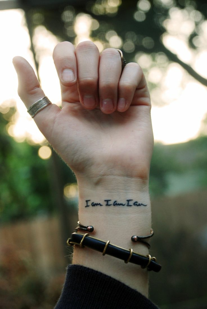 tattoos pequeños, mano con pulsera y anillos, tatuaje en la muñeca con frase en negro cursiva, estilo minimalista