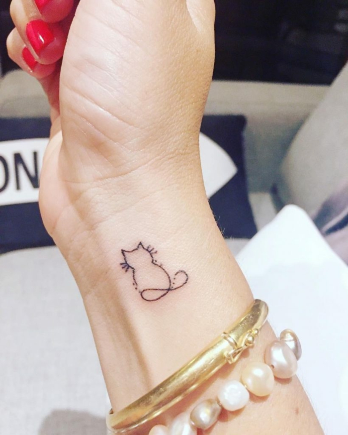 diseño de mini tatuaje con gato estilizado, mano de mujer con uñas rojas y pulseras de oro y perlas, tatuajes en la muñeca