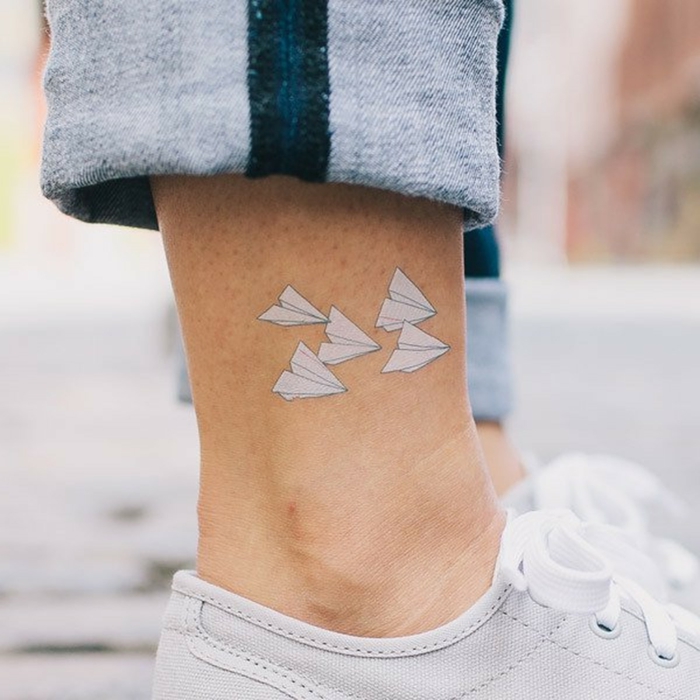 pierna con jeans y zapatillas blancas, tatuaje en el tobillo con aviones origami blancos, tatuajes pequeños con significado