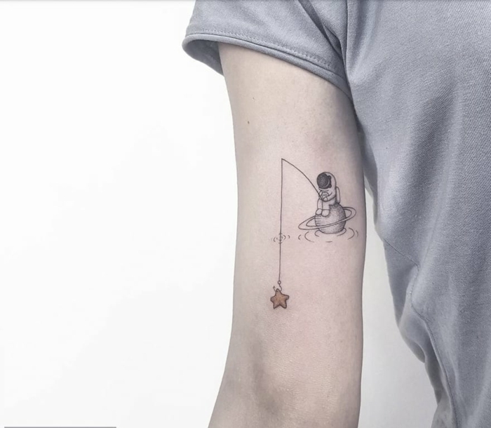 tatuaje divertido en el brazo, blusa gris, astronauta sentado en planeta pescando estrellas, tatuajes simbolicos, diseño original
