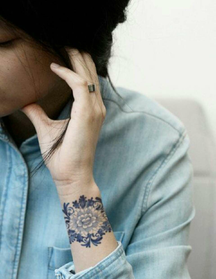 mujer con tatuaje brazalete azul en la muñeca con motivos florales, tatuajes pequeños originales, pelo largo recogido, camisa jeans