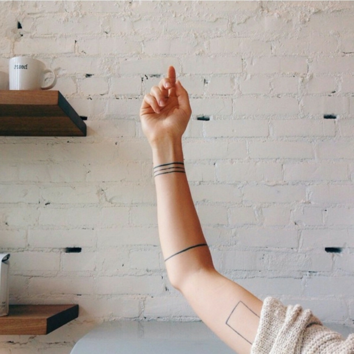 tatuajes en el brazo, brazaletes negras minimalistas, brazo mujer, fondo con pared de ladrillo visto, tatuajes minimalistas