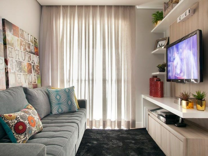decoracion salon pequeño con sofa gris, alfombra negra de pelo, cortinas en color crema y televisor en la pared